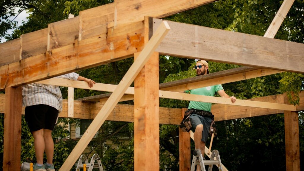 Twee bouwvakkers plaatsen houten constructie op hoogte, werken volgens bouwvoorschriften – wettelijke verplichtingen aannemer
