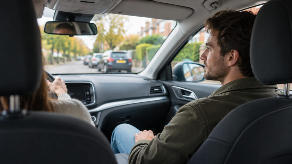 Een coach op de bijrijdersstoel kijkt vanaf de achterbank gezien mee op de weg terwijl de leerling door een rustige woonstraat rijdt