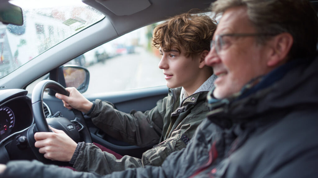 Rijleerling houdt het stuur vast terwijl de rijinstructeur naast hem meekijkt tijdens een rijles